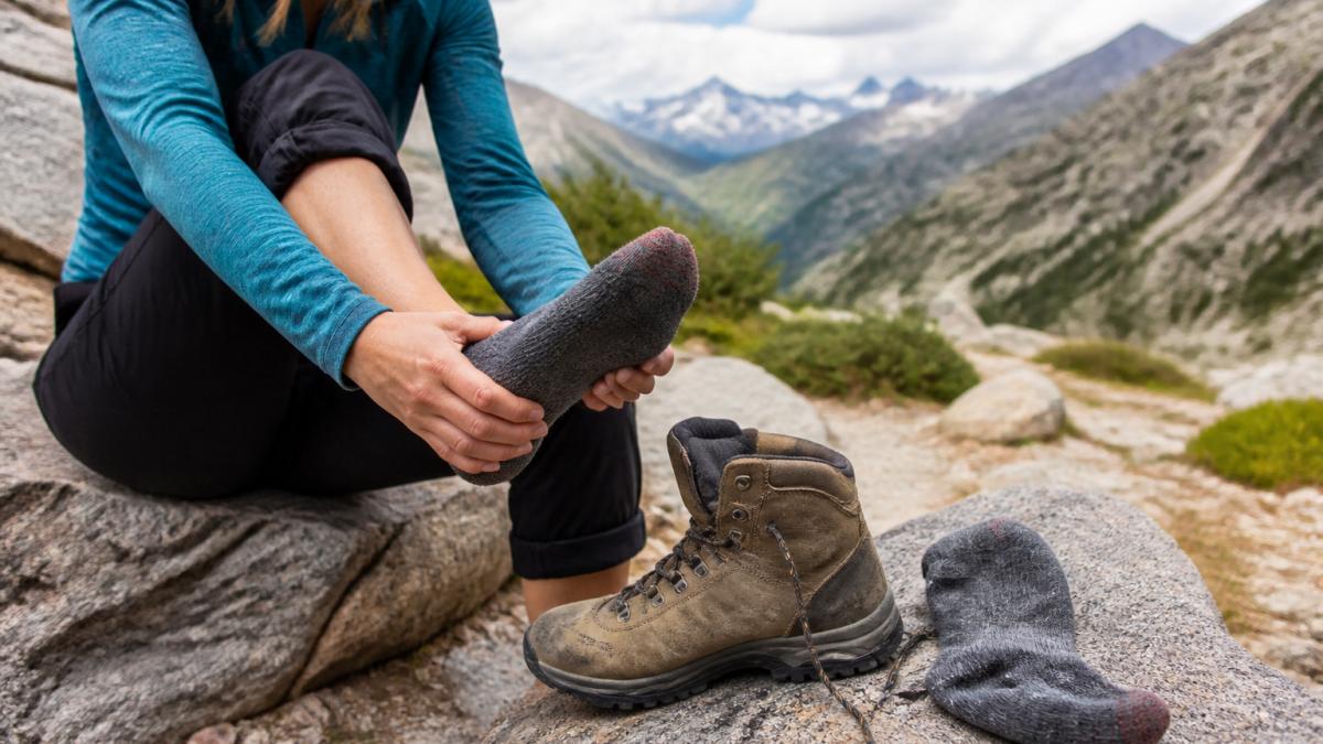 A hiker sitting on a rocky trail pulling off a hiking boot to inspect a sock, with a second sock and boot resting on granite beside them, mountain trail visible in the background
