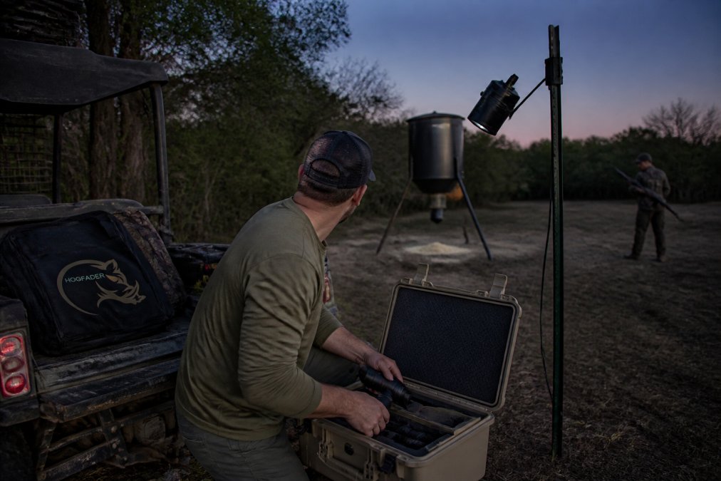 A hog feeder light mounted on a T-post at dusk, angled down toward a feeder in a wooded clearing, with a hunter's silhouette in the background preparing a rifle