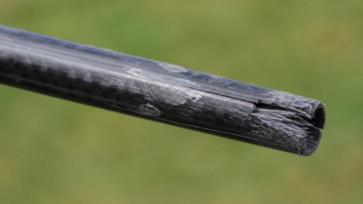 Close-up of a golf shaft showing visible damage including small cracks in graphite material, frayed composite layers at the tip, and surface wear marks under studio lighting