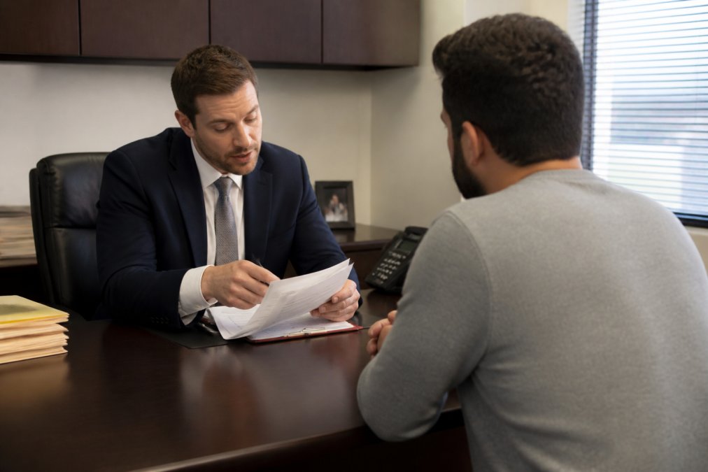 Professional attorney in business attire reviewing documents at a desk with a client seated across, discussing paperwork in a law office setting