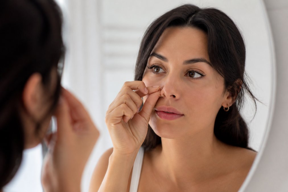 Close-up of a woman's hand gently adjusting a stainless steel clip-on nose ring in a bright bathroom mirror, showing the secure fit on her nostril