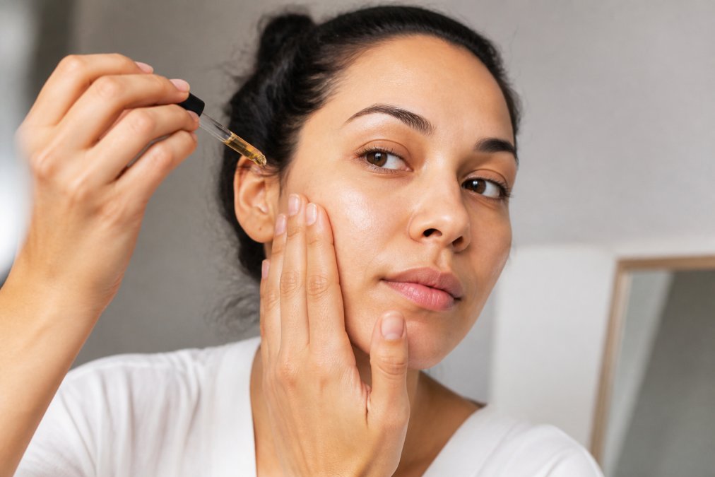 Close-up of hands applying a few drops of facial oil to clean, damp skin on the cheek and forehead, showing proper technique with upward pressing motions and minimal product amount