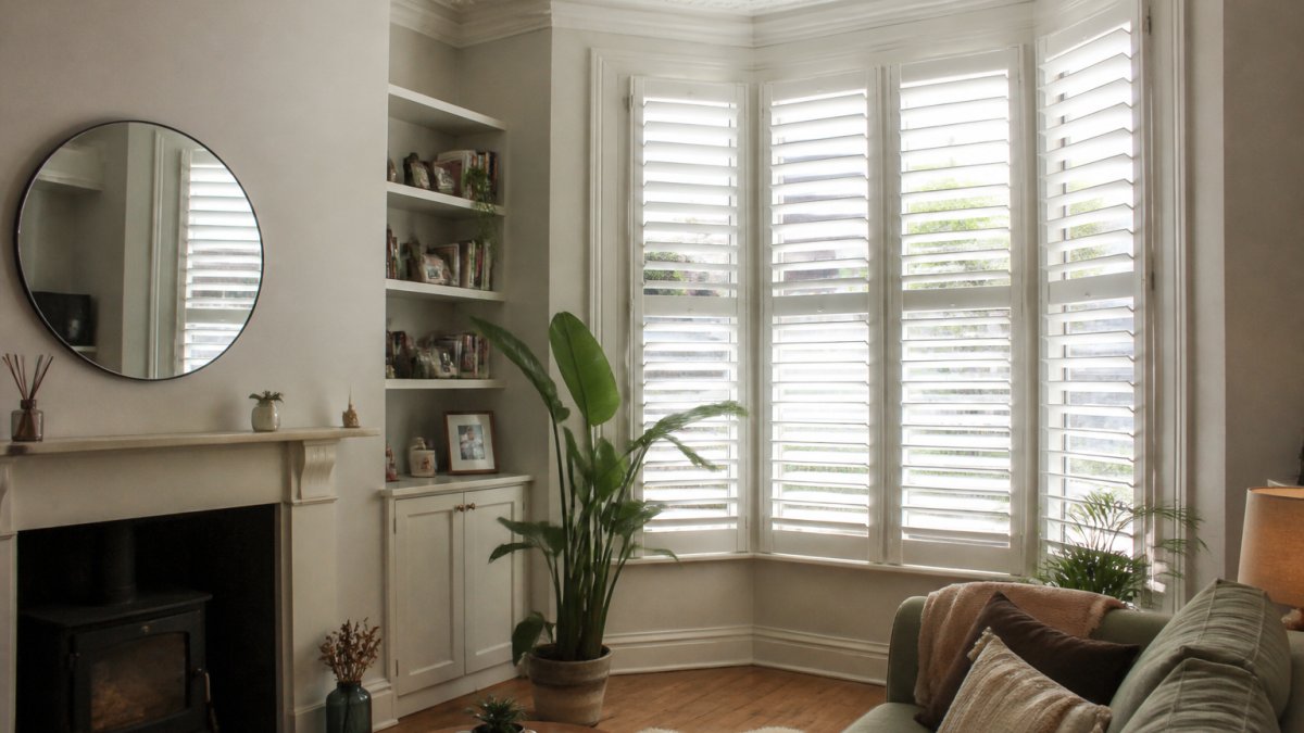 A sunlit Victorian terraced living room with a wide bay window fitted with white full-height plantation shutters, tilted open to let in light, with a sofa and houseplant in view