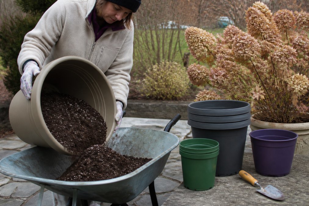 A person in a fleece jacket and garden gloves emptying dark potting soil from a plastic planter into a metal wheelbarrow on a chilly autumn patio, stacked clean pots and a garden trowel nearby, soft overcast afternoon light