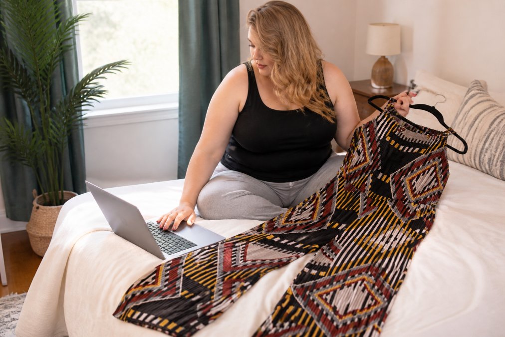 Woman reviewing a plus size jumpsuit while checking a size chart on her laptop, with the garment laid out on a bed with natural window lighting