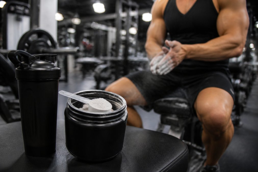 Close-up of a metal scoop resting in a tub of pre-workout powder next to a black shaker bottle on a gym bench, blurred weight room and a lifter chalking hands in the background