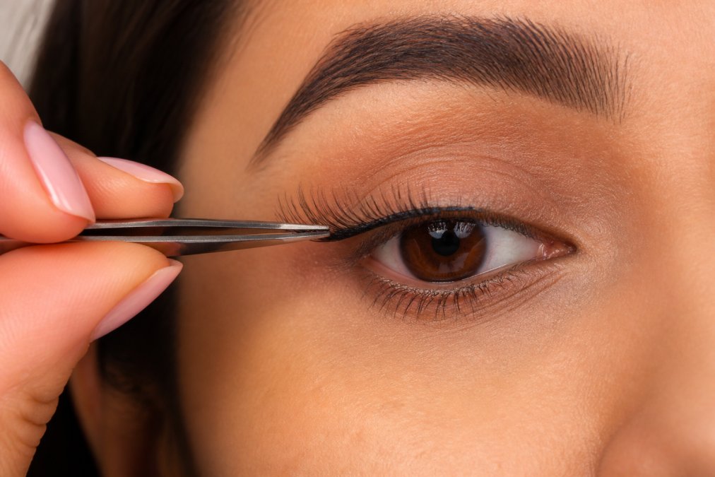 Close-up of hands using precision tweezers to position a lash cluster at the natural lash line, showing proper under-lash placement technique with clear visibility of tapered fibers
