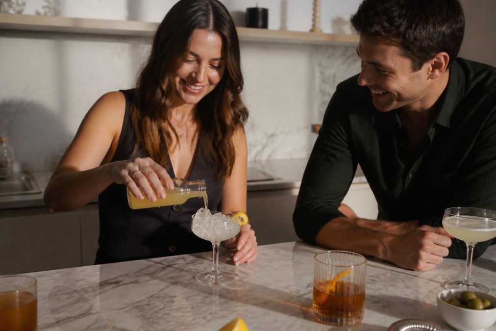 A person pouring a chilled ready-to-drink cocktail from a small glass bottle into a coupe glass filled with ice, with a fresh lemon twist garnish resting on the rim, on a marble kitchen countertop in soft afternoon light