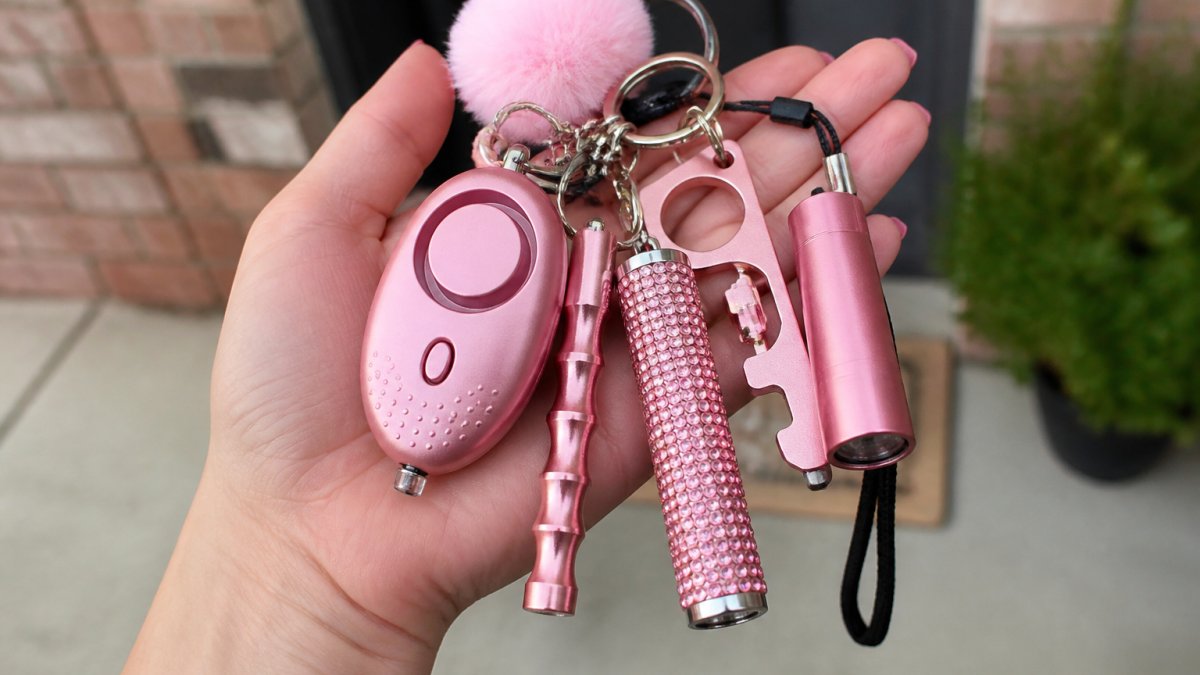Close-up of a woman's hand holding an organized safety keychain set with multiple tools visible, including alarm, flashlight, and other accessories, displayed against a neutral background