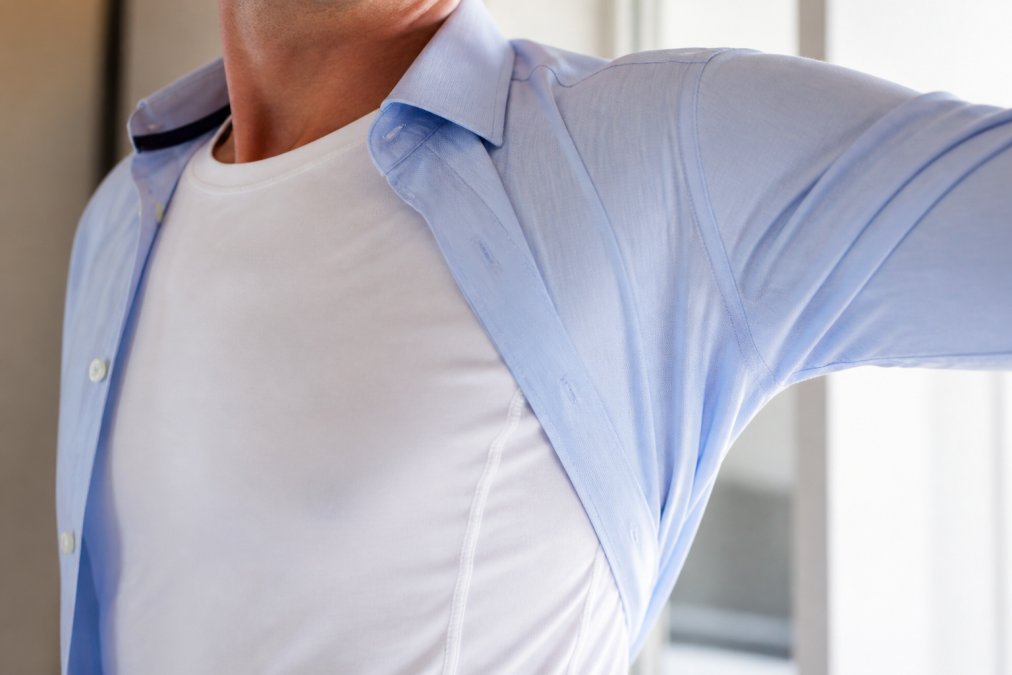 Close-up of a person's torso wearing a white undershirt under an unbuttoned dress shirt, showing the fitted construction and underarm area in a bright, neutral setting