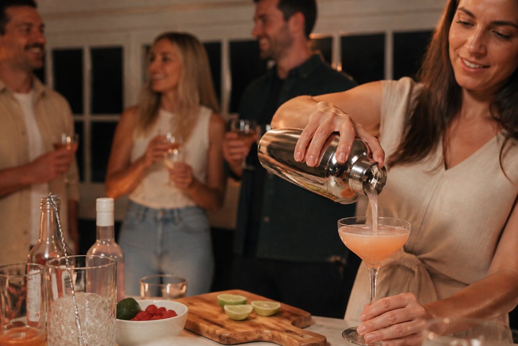 Someone pouring a freshly made cocktail into a glass at a home gathering, with guests in the background enjoying drinks
