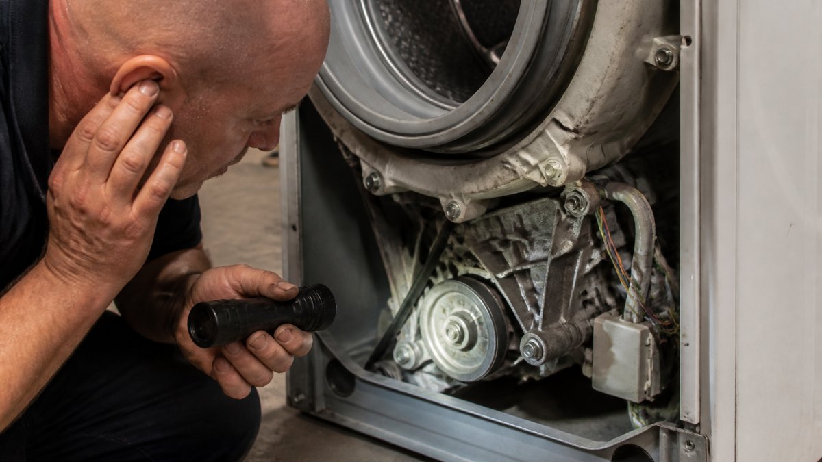Close-up of a technician's hands holding a torch beside an open front-loader washing machine, listening to a worn gearbox coupling, workshop lighting