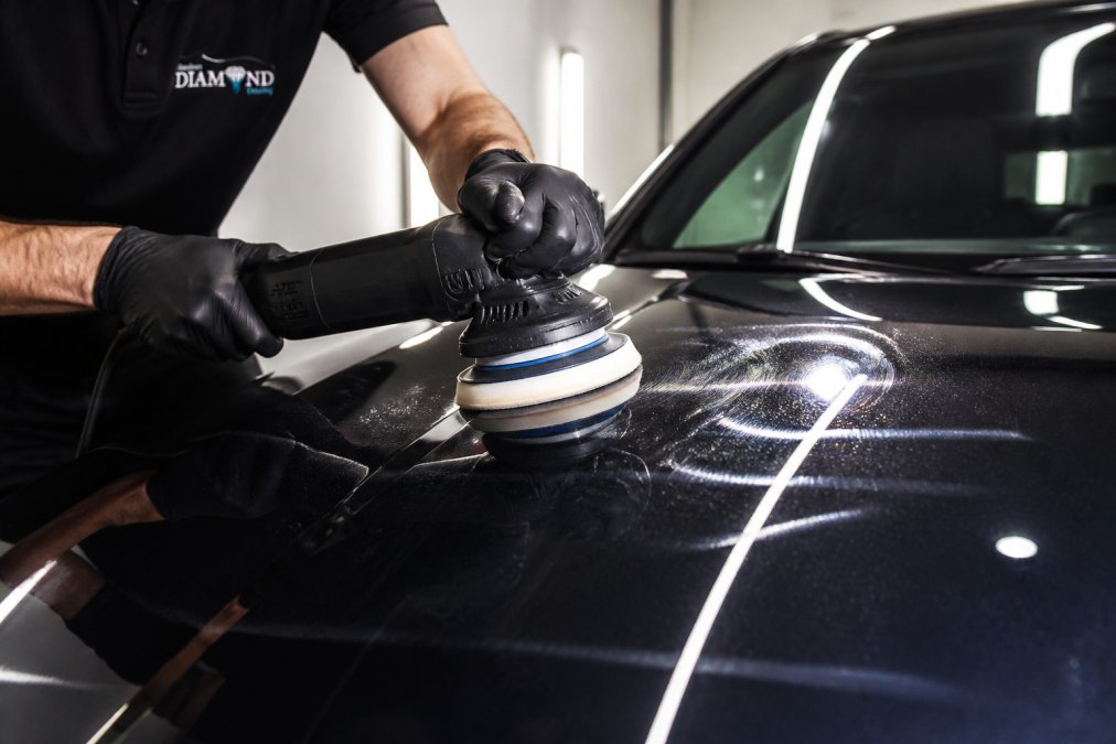 Detailer using a dual-action machine for paint correction on a dark prestige car bonnet under bright workshop lighting.