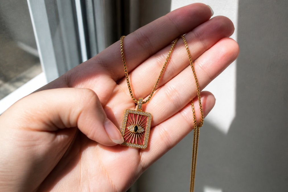 Close-up of a woman's hand holding an 18K gold-plated necklace, showing slight surface discolouration and tarnish against natural daylight streaming through a window