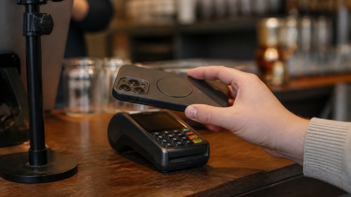 Close-up of a hand holding a smartphone with an E Ink phone case to a contactless payment terminal at a cafe counter, warm indoor lighting, shallow depth of field