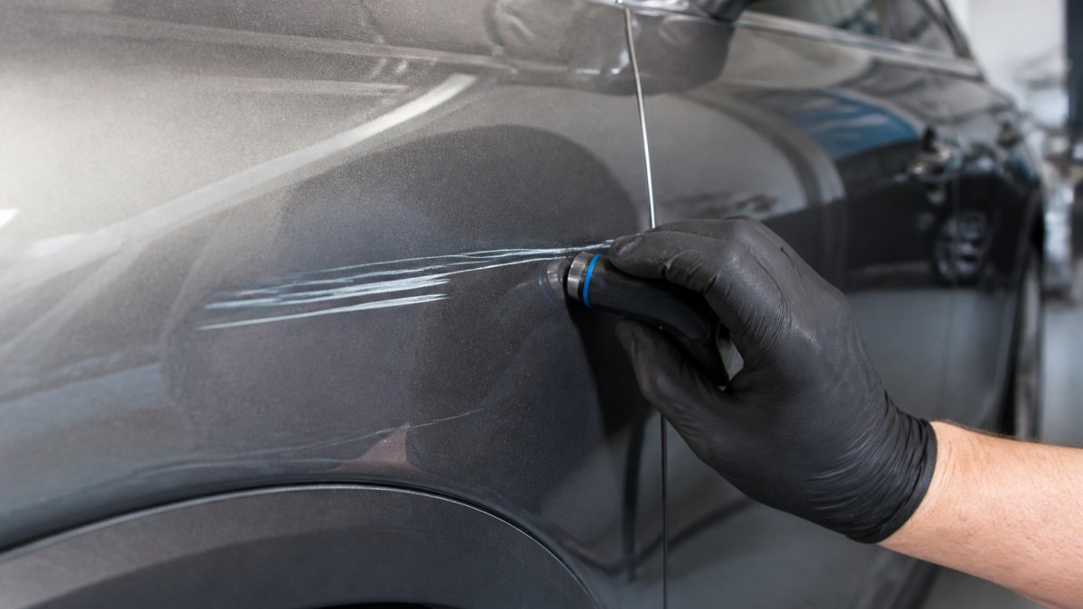Close-up of a vehicle's side panel showing a deep scratch through the clear coat, with a technician's gloved hand holding a paint depth gauge near the damaged area in a well-lit auto body shop