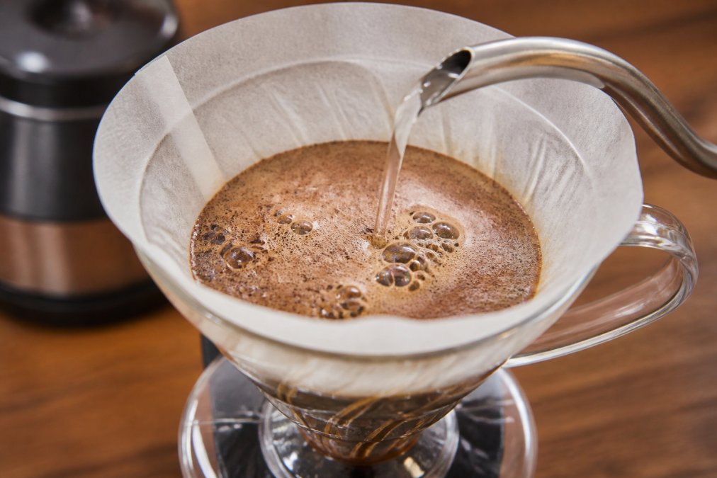Close-up of water being poured slowly over coffee grounds in a pour-over dripper, showing visible bubbling and gas release from the coffee bed during the bloom phase
