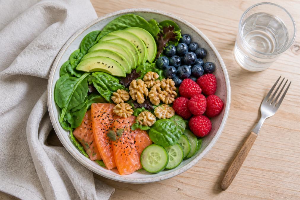 Overhead shot of a colourful bowl of salmon, leafy greens, walnuts, avocado and berries on a light timber table, with a glass of water nearby