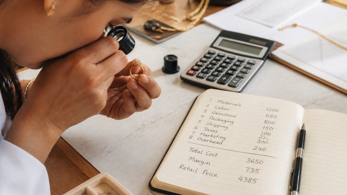 A close-up of a jeweler's hands examining a gold ring with a loupe on a clean workbench, with a notebook showing handwritten cost breakdown notes nearby