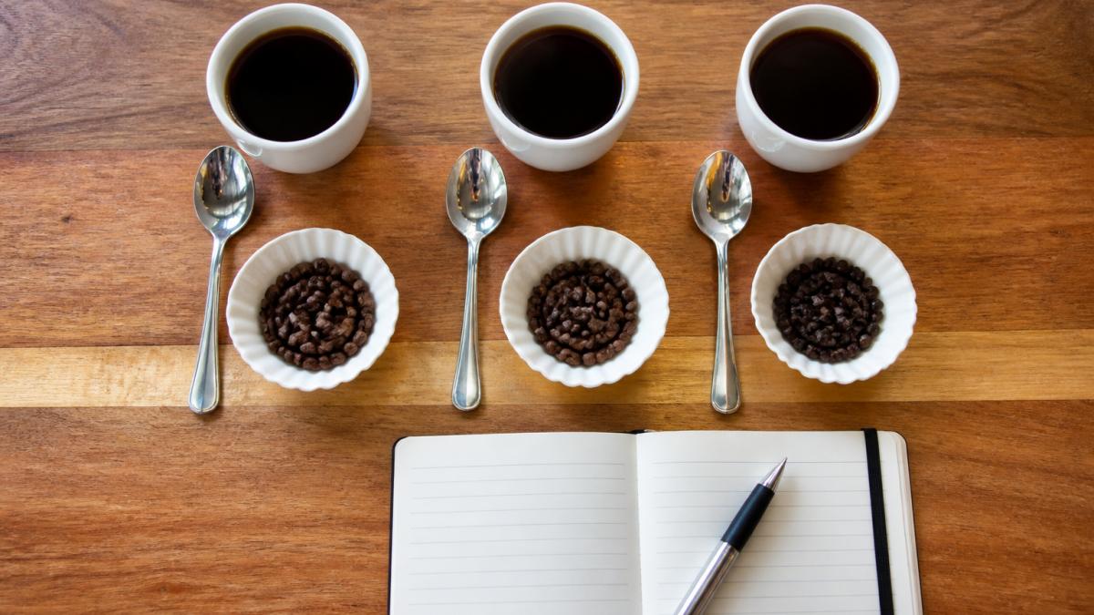 Overhead shot of three small white cups of black coffee on a wooden table, each beside a spoon and a small dish of coffee beans, with a notebook and pen nearby for tasting notes