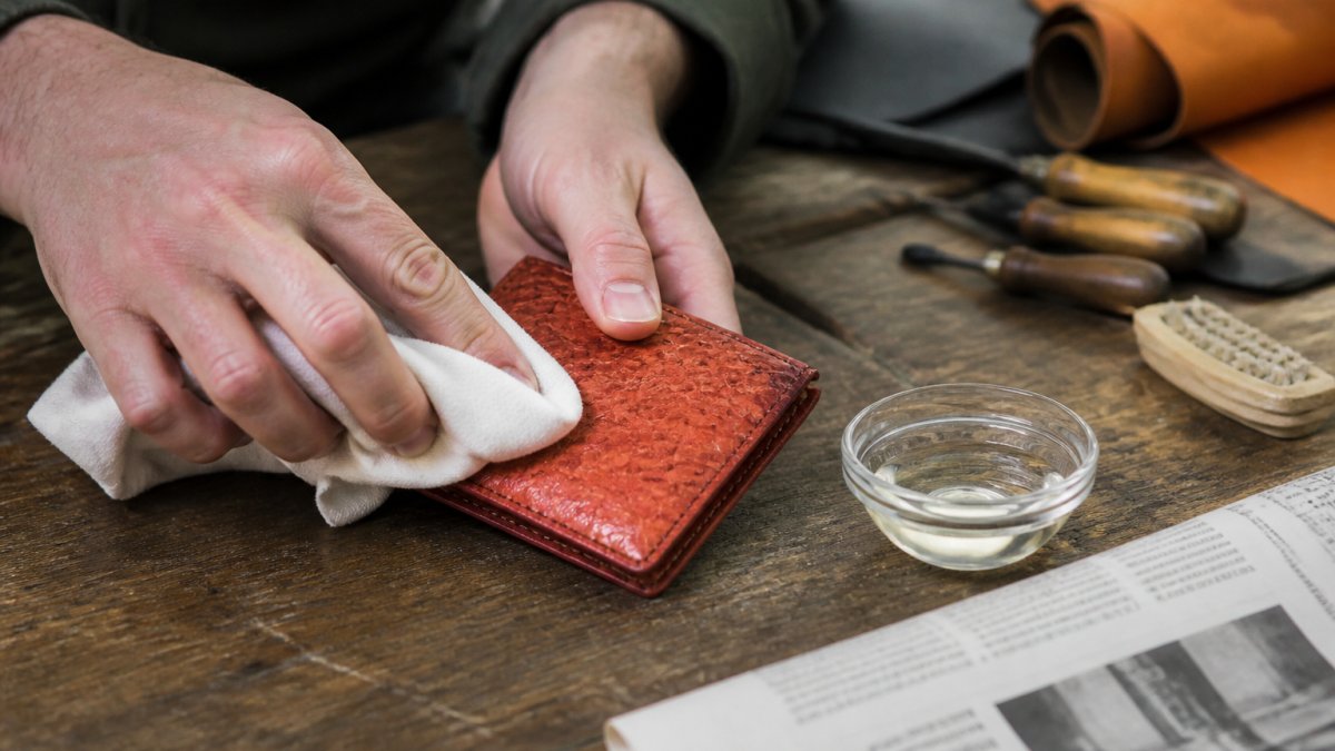 Close-up of hands gently wiping a salmon leather wallet with a soft white cloth on a wooden workbench, with a small bowl of diluted white vinegar and a sheet of newspaper nearby