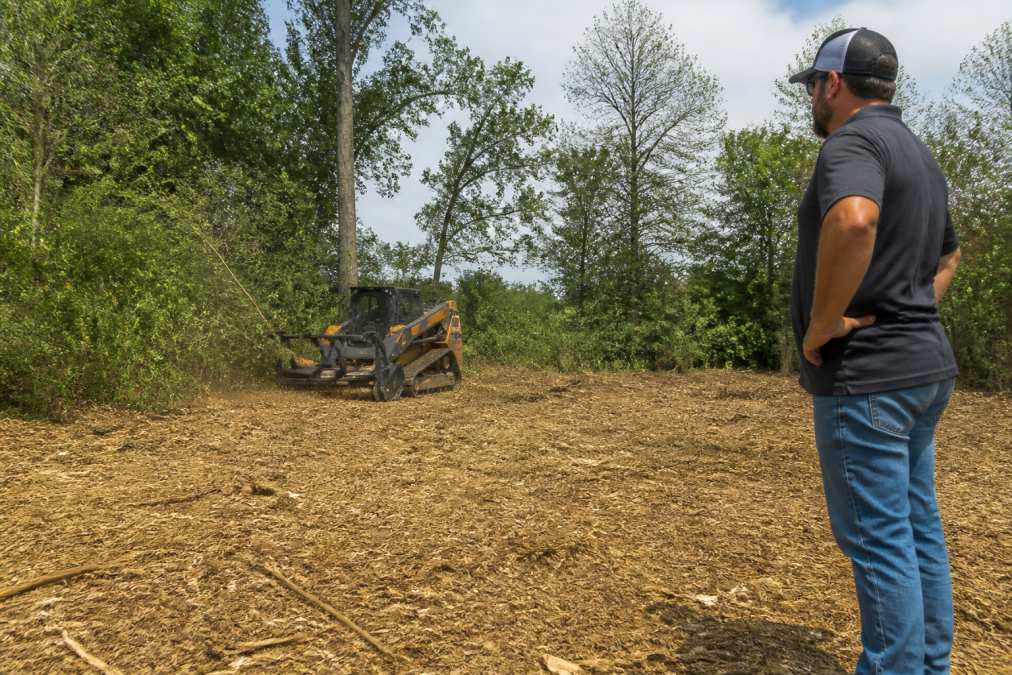 A forestry mulching machine clearing thick brush on a wooded property, with freshly cut mulch spread across the ground and mature trees in the background