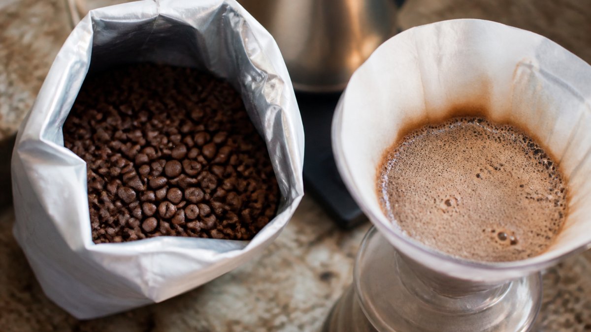 Close-up of freshly roasted whole coffee beans in an open bag on a kitchen counter, with a small bloom of bubbles visible on a pour-over cone nearby
