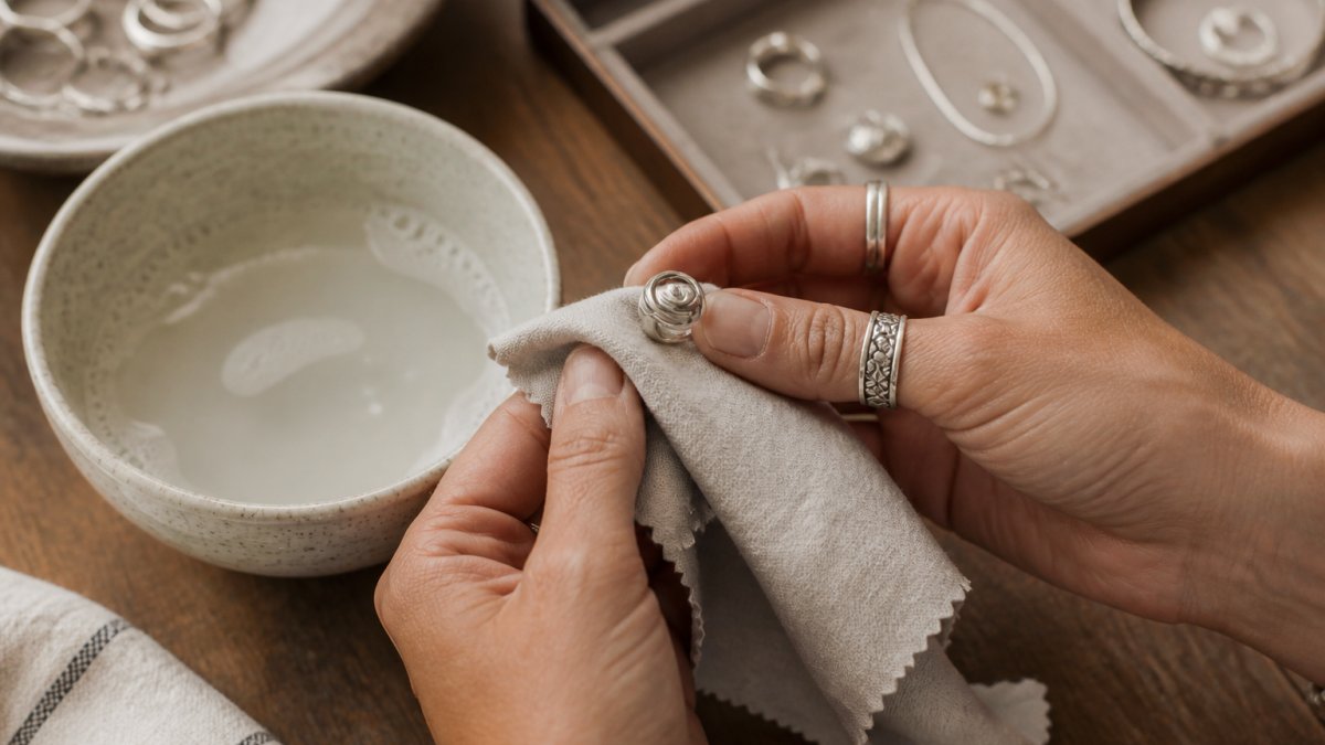 Close-up of hands gently polishing a handmade silver ring with a soft cloth on a wooden table, with a small bowl of cleaning solution and a jewelry box nearby