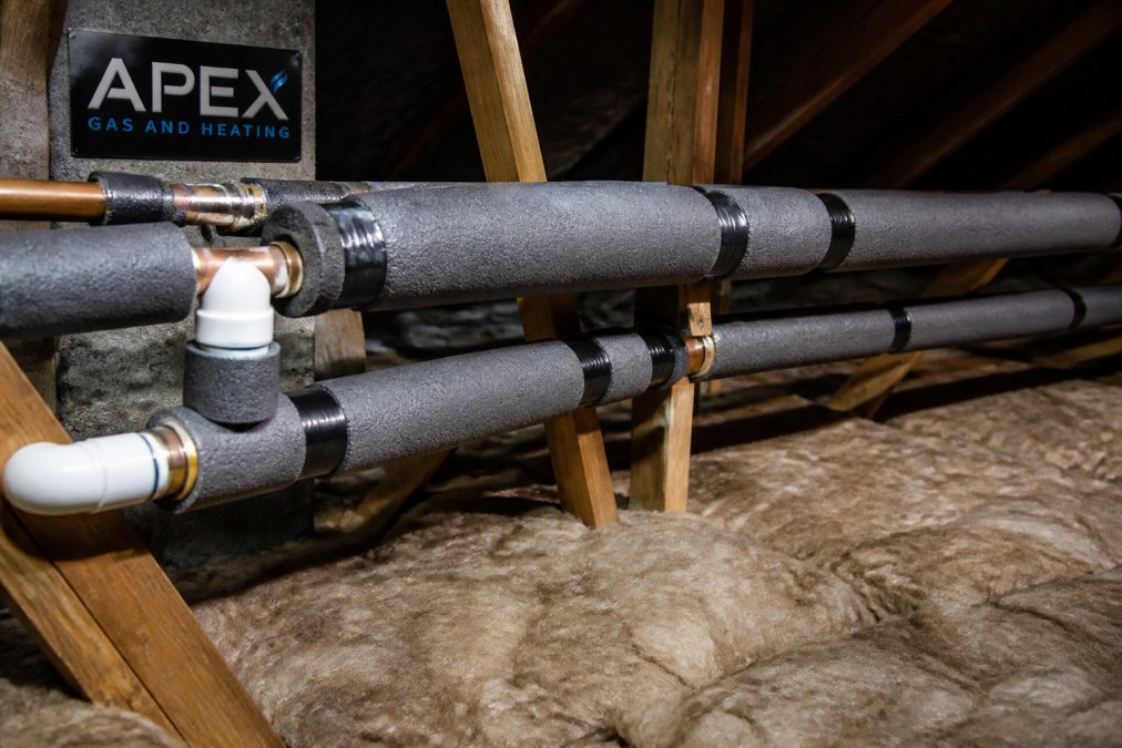 Exposed copper and plastic pipes wrapped tightly with thick foam pipe insulation in a cold, unheated loft space, showing proper installation technique with insulation secured with tape
