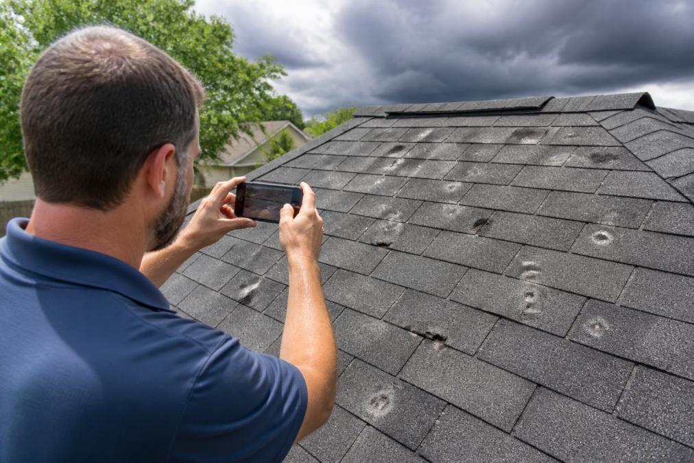 Homeowner photographing roof damage with smartphone, showing visible hail dents and water stains on asphalt shingles in bright daylight, with storm clouds in the background