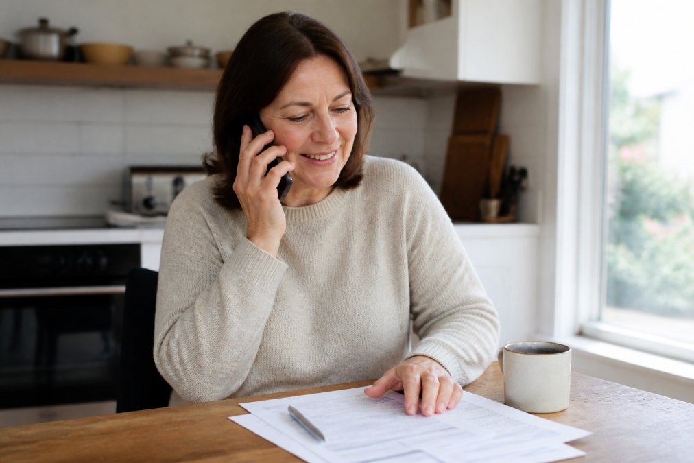 A person sitting at a kitchen table with a smartphone, insurance paperwork, and a cup of tea, looking relieved while making a call after a car accident