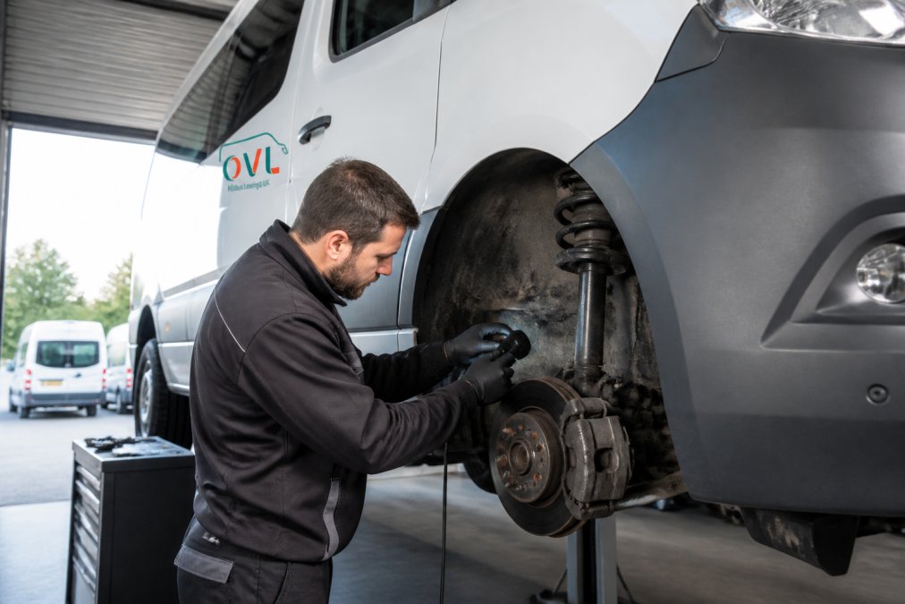 Professional technician performing systematic safety inspection on modern minibus in bright workshop, checking brake systems and suspension components with diagnostic equipment