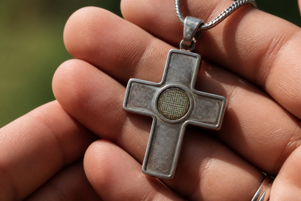 Close-up of a nano-engraved cross pendant necklace held in hands with natural lighting, showing a Bible verse chip with subtle oxidation or surface discoloration on the engraved area and surrounding metal