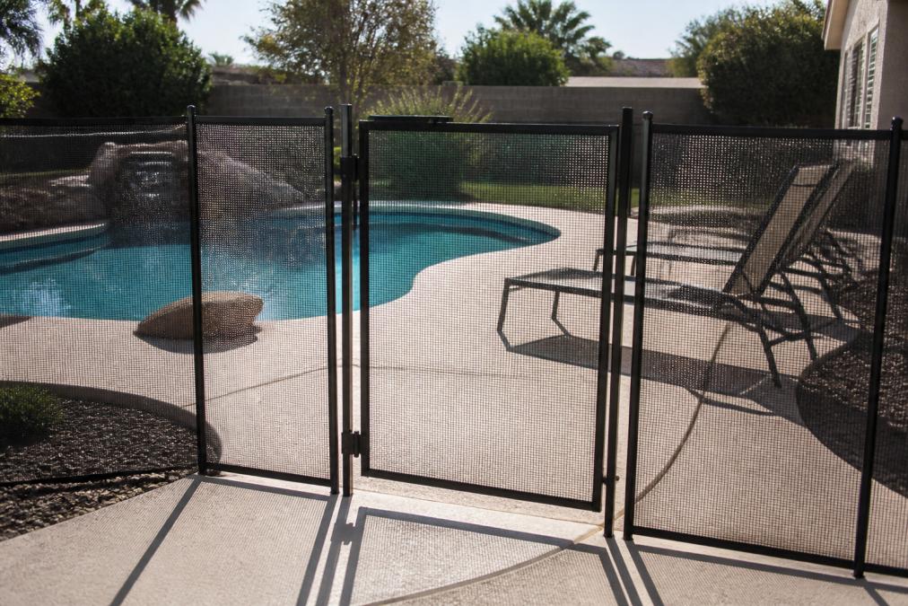 Residential backyard with properly installed safety fence around in-ground pool, self-closing locking gate clearly visible, afternoon sunlight casting shadows on the pool deck