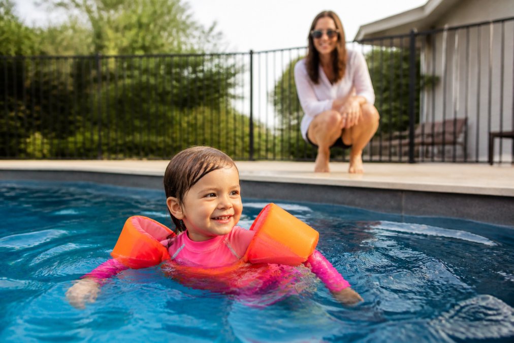 Young child wearing bright high-visibility swimwear and rash guard in pool setting with clear view of parent supervising from pool deck, natural daylight