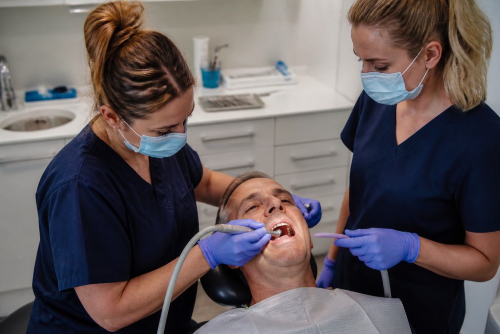 A dental hygienist in a bright modern treatment room using an ultrasonic scaler on a patient's lower teeth, with a dental nurse assisting and professional equipment visible in the background