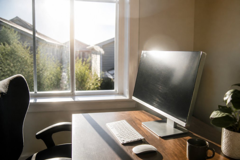 Bright home office with afternoon sunlight streaming through untinted windows, creating visible glare on a computer monitor and desk surface, showing the problem window tint solves