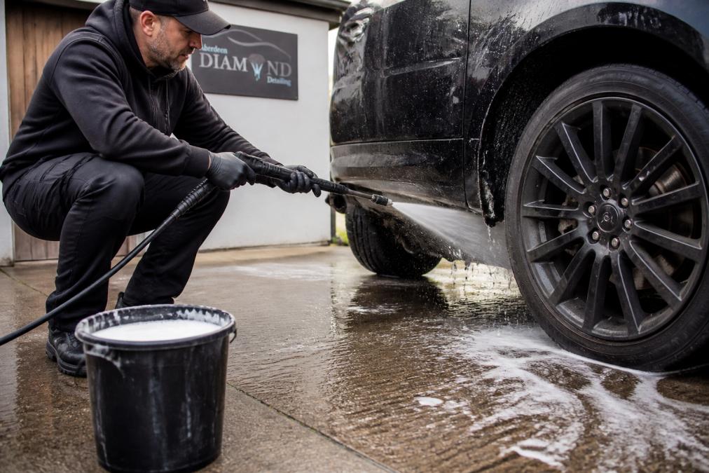 Close-up of a person using a pressure washer to rinse the underside of a car, water spraying across the wheel arch, bucket of soapy water on the driveway beside them