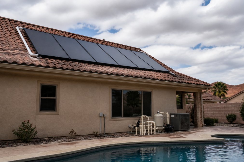 Rooftop solar thermal collector panels installed on a residential home under a partly cloudy sky, with visible pool equipment and plumbing connections nearby