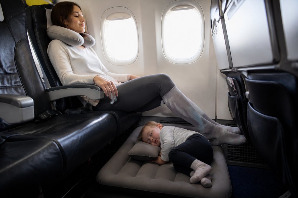 A parent sitting in an airplane seat with a toddler asleep on an inflatable travel bed placed on the floor, while the parent wears compression socks and a neck pillow, cabin interior visible