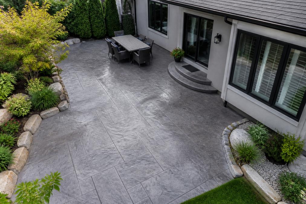 Overhead view of a finished stamped concrete patio with decorative slate pattern adjacent to a modern home exterior showing landscaping and manicured curb appeal