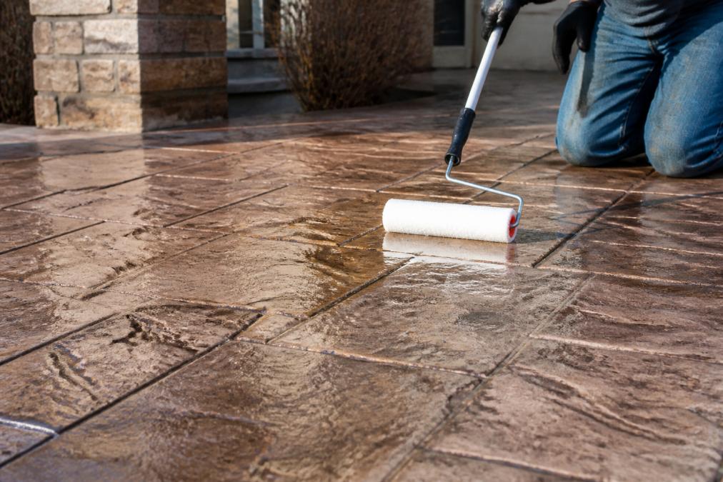 Close-up of a homeowner's hands applying a clear protective sealant to stamped concrete surface with a roller, showing the decorative texture and professional care being applied in natural daylight