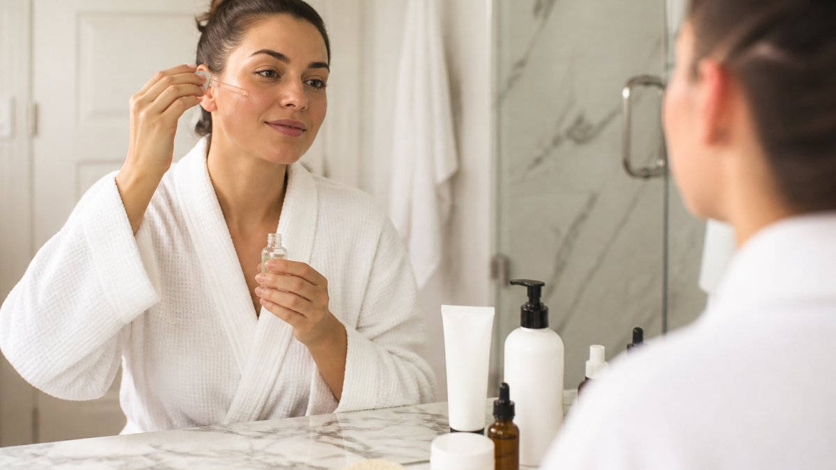Woman applying clear vitamin C serum from a glass dropper to her face in a bright bathroom setting