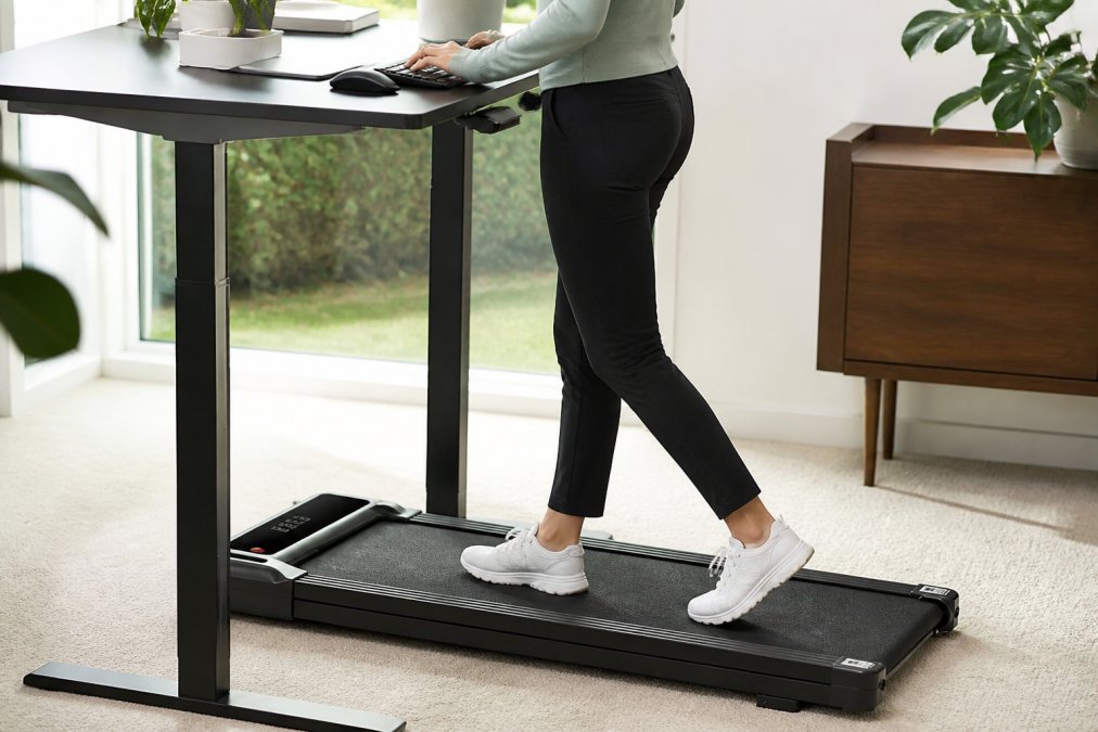 Close-up of a person's hands on a keyboard with a foldable walking treadmill visible beneath the desk, demonstrating proper ergonomic positioning and desk setup with natural office lighting