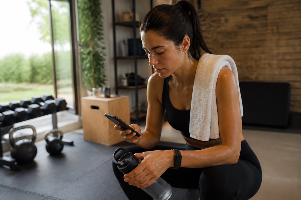 Person reviewing fitness tracker data on smartphone while holding a water bottle after a workout session in a home gym setting