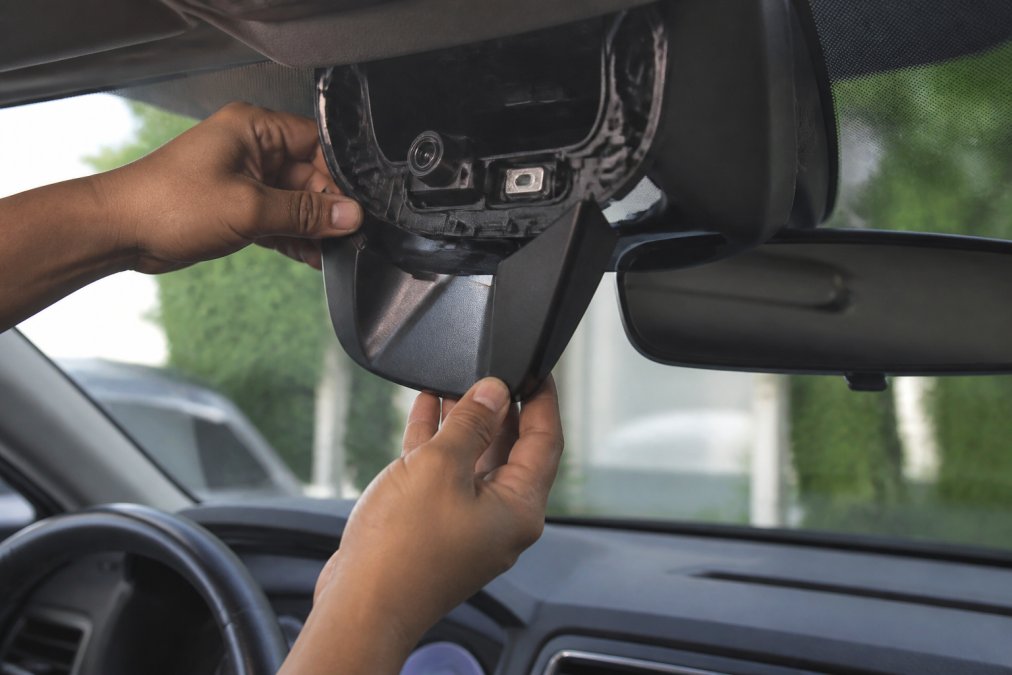Close-up of a technician's hands carefully removing the plastic cover around a windshield-mounted camera on a modern car interior, with the rearview mirror and dashboard visible in soft daylight