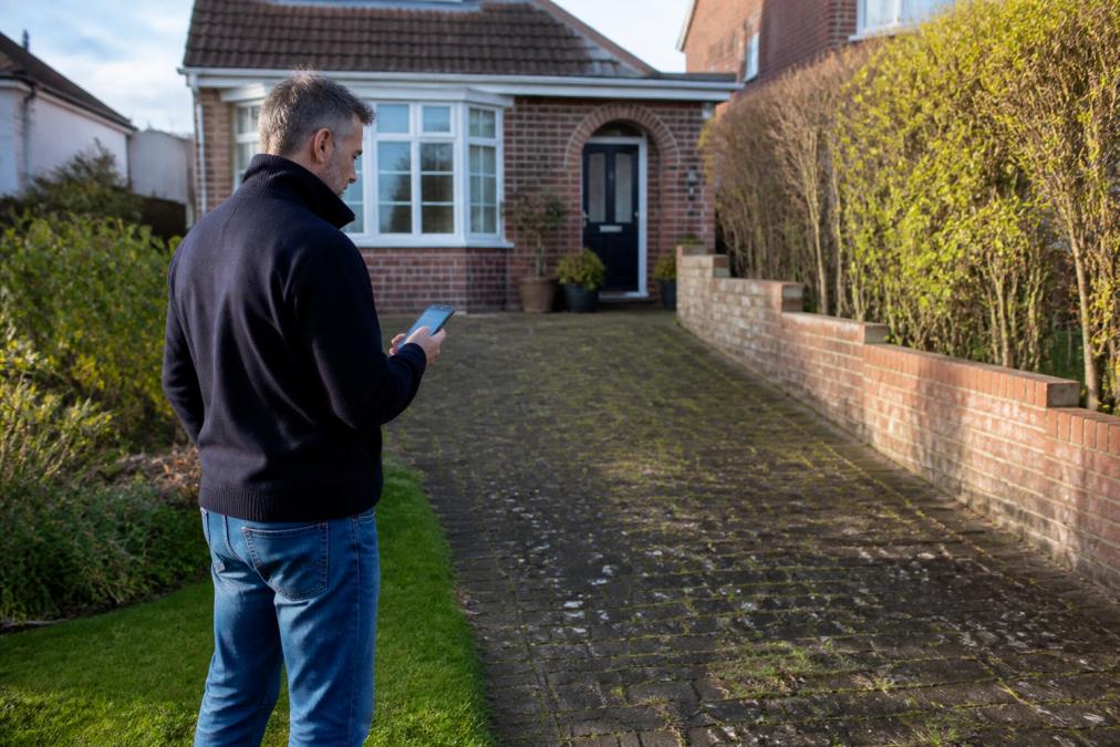 A homeowner in Norfolk standing in their front garden, holding a smartphone while examining the existing driveway surface and front boundary
