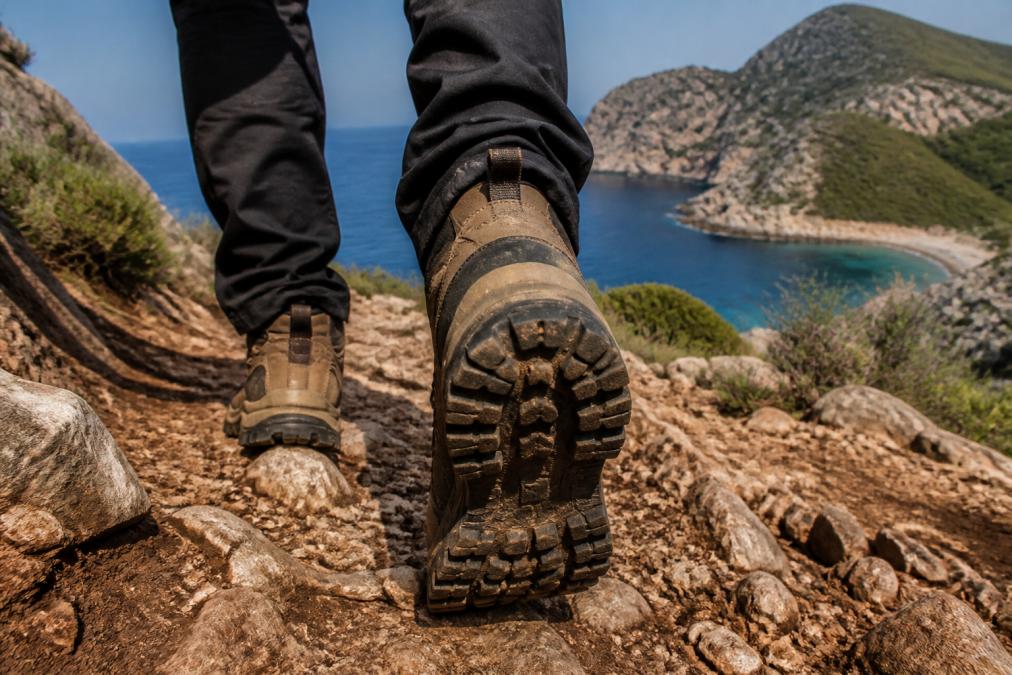 A close-up of a hiker's feet wearing professional hiking boots with thick treads, stepping over rocky terrain on a coastal trail in Fethiye, with the Mediterranean Sea visible in the background