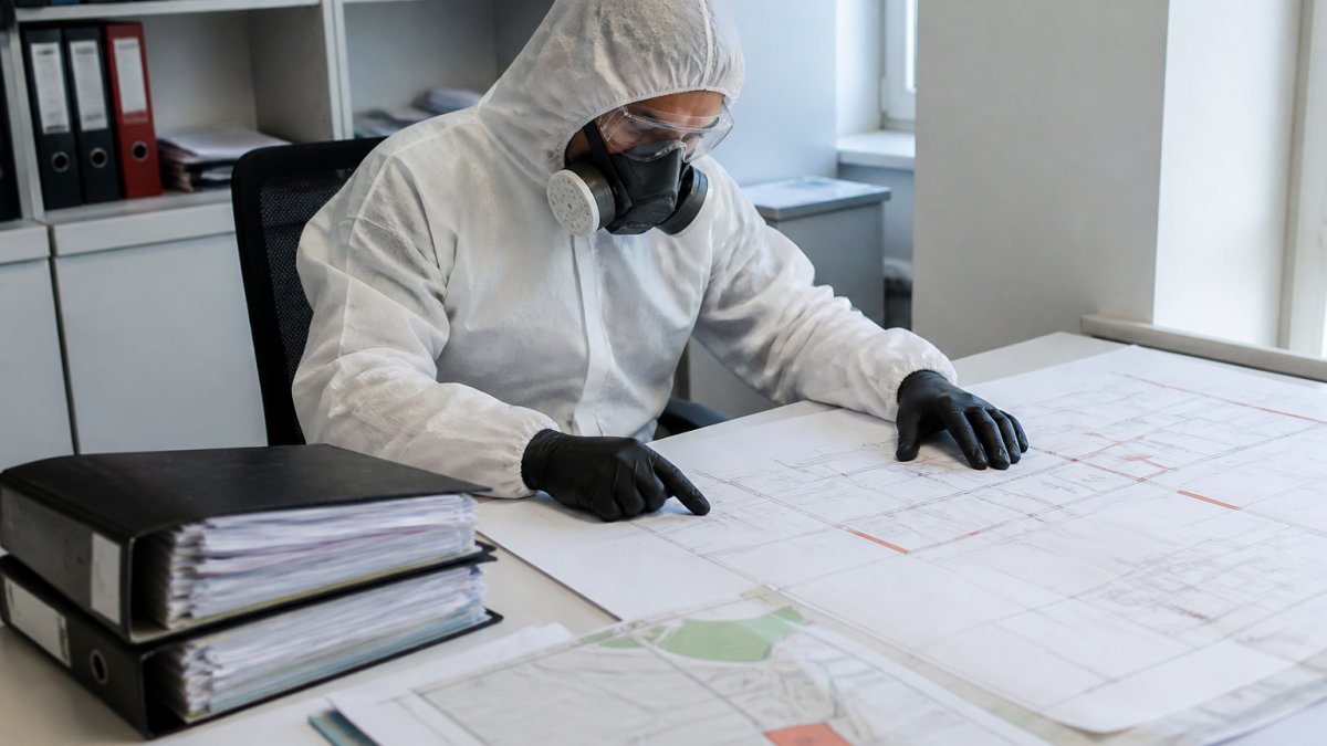Certifikovaný pracovník v bílém ochranném obleku a respirátoru pracující se schematickými plány budovy na stole, s dokumenty a mapami rozložení azbestových materiálů v moderní kanceláři