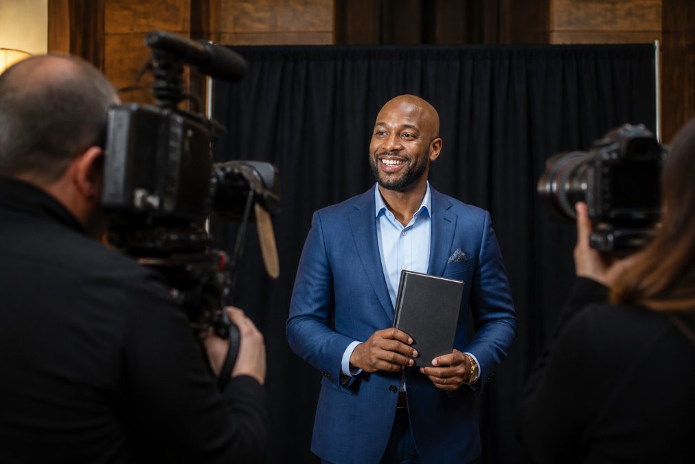 A business professional in a tailored suit holding a published hardcover book while standing confidently in front of a media backdrop with studio lighting, conveying a TV or press interview setting
